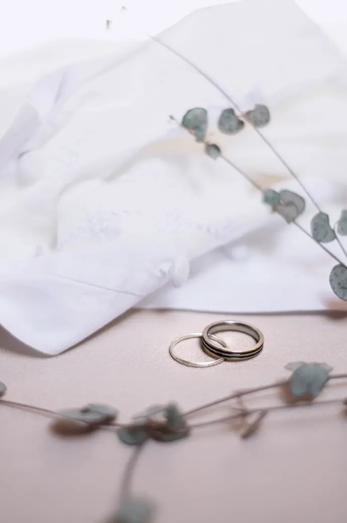 alliances posées sur un tissu beige devant la robe de mariée et avec des feuilles décoratives disposées autour - photo Urielle PENN photographe de mariage